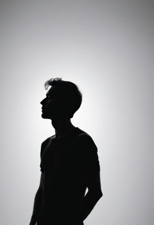 A man stands silently in profile, illuminated by a bright light from behind, casting a striking silhouette that evokes feelings of reflection and tranquility in a minimalist setting.の素材