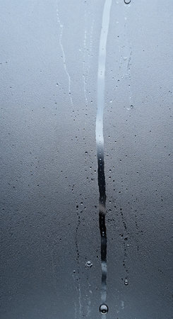 An abstract view of a wet glass surface featuring many small water beads and a distinct, thick stream flowing downwards, creating a dynamic wet weather pattern.の素材