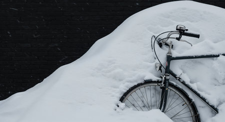 A close-up view reveals a dark-framed bicycle partially submerged in a mound of soft white snow, contrasting sharply with the dark, textured brick background.の素材
