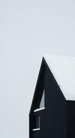 The striking side of a contemporary dark house, featuring a steep gabled roof laden with pristine snow and prominent triangular and rectangular windows, stands out under a bright, clear winter sky.の素材