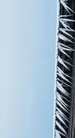 Numerous long, pointed icicles with accumulated snow adorn the rustic wooden eaves of a structure, dramatically contrasting with the expansive, serene light blue sky on a cold, crisp day.の素材