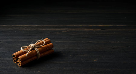 Several aromatic cinnamon quills, neatly bound together with a simple rope, are prominently displayed on a deeply textured black wooden background, offering a cozy, warm aesthetic.の素材