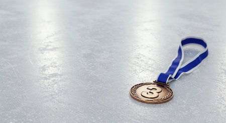 The third-place bronze medal with its distinctive blue and white ribbon is carefully placed on a brightly lit, scratched ice rink, symbolizing achievement and competition.の素材