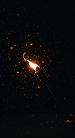 A vibrant orange torch flame, held by a person's hand, emits a shower of sparkling light particles against the profound darkness of a mysterious night.の素材