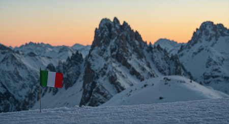 A breathtaking winter landscape showcases a small Italian flag planted in the foreground, with towering, dramatic peaks and a soft, colorful twilight glow across the horizon.の素材