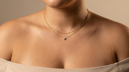 Close-up of a person wearing an elegant off-the-shoulder beige top, showing a beautiful gold chain necklace with a single brilliant diamond on their smooth skin.の素材