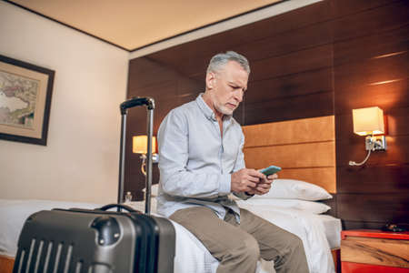 Mid-aged businessman sitting on a bed in a hotel with a smartphone in handsの写真素材