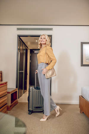 Elegant blonde woman in grey pants standing in a hotel roomの写真素材