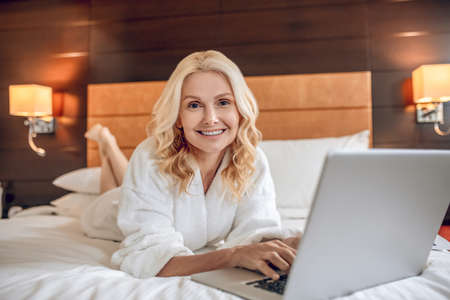 Pretty woman in a bath robe laying on bed with a laptop and typinga messageの写真素材