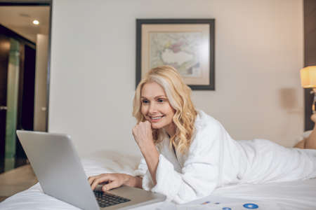 Pretty woman in a bath robe laying on bed with a laptop and typinga messageの写真素材