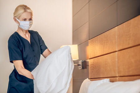 Blonde woman in a protective mask making bed in a hotel roomの写真素材