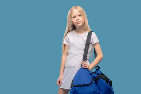 Hobby, sports. Blonde calm girl in white suit with sports bag standing on blue background in good moodの写真素材