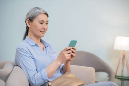 Mature good-looking woman sitting in the chair with a smartphone in handsの写真素材