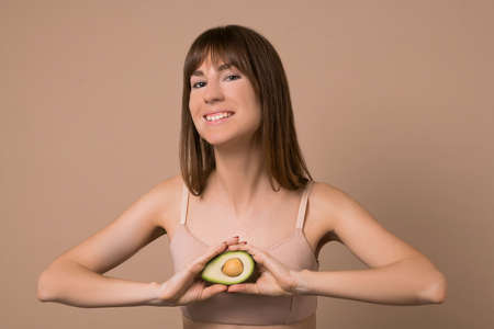 A slim long-haired woman in beige lingerie with avocado in her handsの写真素材