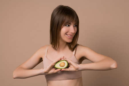 A slim long-haired woman in beige lingerie with avocado in her handsの写真素材