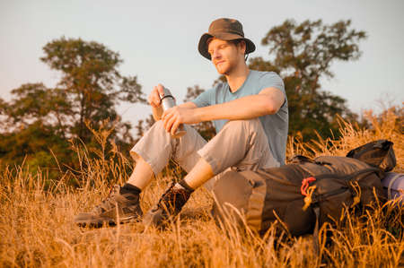 A man in a hat sitting on the hill and opening his flaskの写真素材