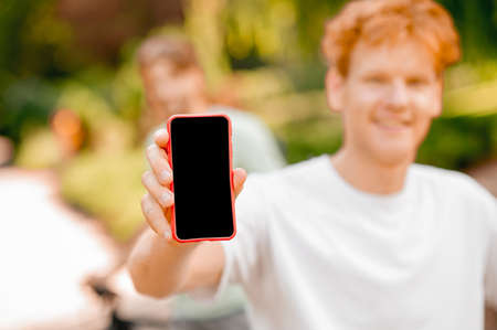 Pleased ginger-haired guy demonstrating his cellular phone outdoorsの写真素材