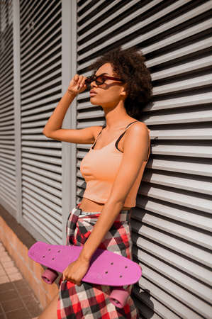 A young mulatta with a skateboard in hands standing near the wallの写真素材