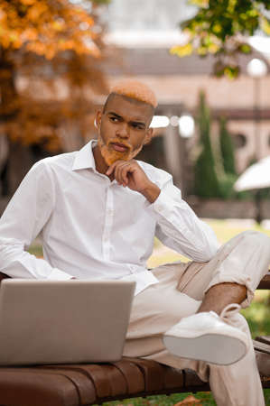 A young man with dyed hair sitting on the bench in the park and working on laptopの写真素材
