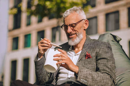 Pleased bearded gray-haired male eating food with chopsticks outside
