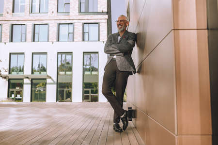 Cheerful bespectacled gray-haired bearded standing before the office buildingの写真素材
