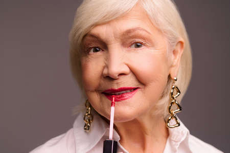 Blonde stylish senior woman putting lip gloss on her lipsの写真素材