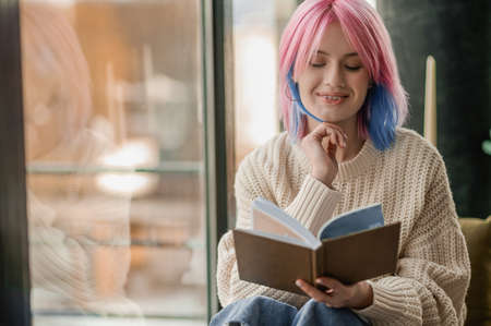 A girl with pink hair reading a book near the windowの写真素材