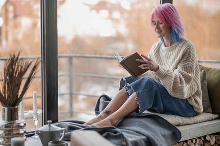 A girl with pink hair reading a book near the windowの写真素材