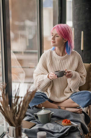 Young girl with pink hair having tea near the windowの写真素材