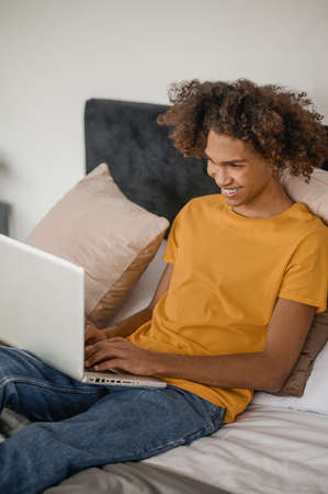 Curly-haired young guy sitting on a bed with a laptopの写真素材