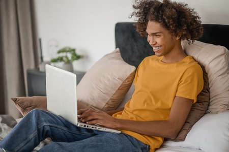 Curly-haired young guy sitting on a bed with a laptopの写真素材