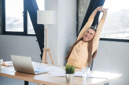 A man in casual clothes sitting at the table in the office and stretchingの写真素材