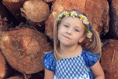 A 3 years old girl with chamomile wreath is standing in front of a wood stackの写真素材