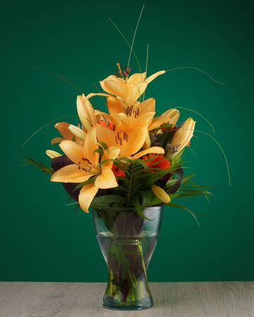 A bunch of yellow lily flowers in a glass vase on a table against a green wallの写真素材