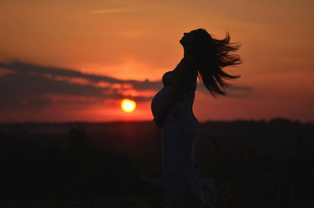 Young pregnant woman stands in the grass at sunset. Pregnant woman in dress standing in the sunset. Silhouette of a pregnant woman on the sky background Silhouette of a pregnant woman at sunsetの写真素材