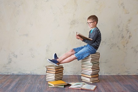 The boy student in sneakers shorts and a sweater sitting on a stack of books and reading. On a yellow colored background. Educational conceptの写真素材