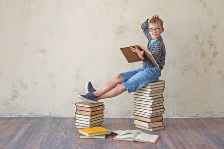 The boy student in sneakers shorts and a sweater sitting on a stack of books and reading. On a yellow colored background. Educational conceptの写真素材
