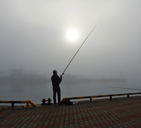 A full-length fisherman with a fishing rod in the fog on the pier. Galloの写真素材