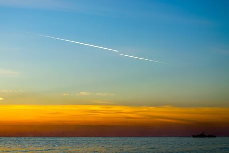Contrails of aircraft in the blue sky at sunset, sea with ship silhouette on the horizonの写真素材