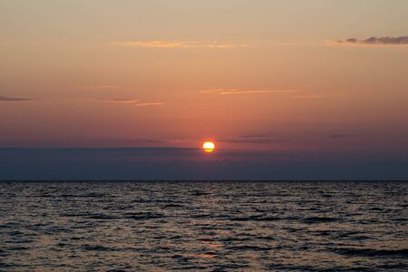 The bright red sun sets over the sea horizon. The sun in the horizontal clouds. The concept of a calm end to a hard dayの写真素材