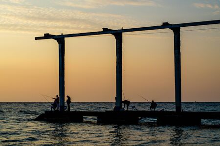 Sochi, Russia - October 19, 2019: Dark silhouettes of fish with fishing rods on a breakwater. A berth for lifting yachts to the shore. Men on vacation. Sea at sunset. The sky is warmの写真素材