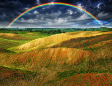 Dramatic sky with a vibrant rainbow over vast, wavy plowed fields on rolling hillsidesの写真素材