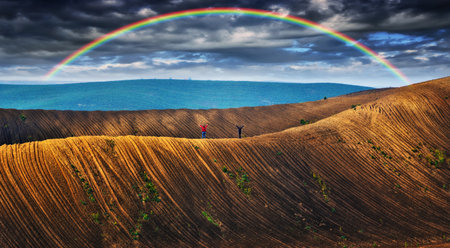 Dramatic sky with a vibrant rainbow over vast, wavy plowed fields on rolling hillsidesの写真素材