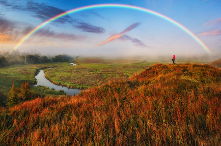 A vibrant rainbow arches over a scenic meadow and river on a clear autumn morningの写真素材