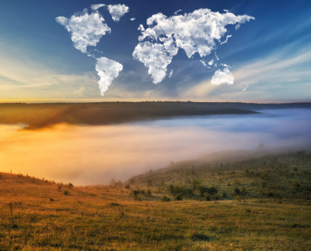Conceptual landscape: cumulus clouds shaped like a world map float over a meadow on a clear morningの写真素材