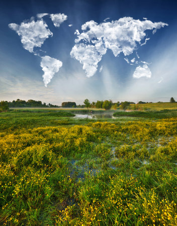 Conceptual landscape: cumulus clouds shaped like a world map float over a green meadow on a clear morningの写真素材