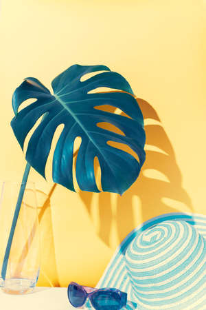 Monstera leaf in a glass vase a straw hat on a yellow background. Tourism and travel conceptの写真素材