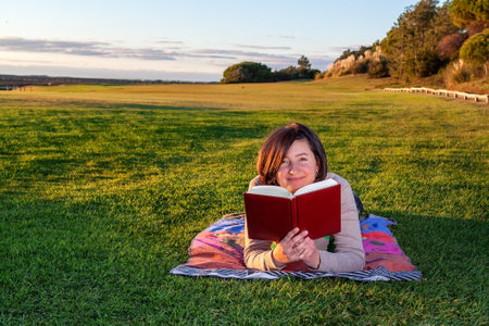 Woman lying down on some grass, smiling while holding a red book and looking away. High quality photoの写真素材