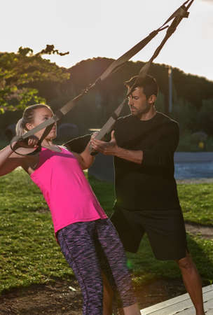 Personal fitness trainer and young woman client, corrects exercises with straps. In the park in the...