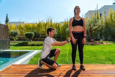 A European man, a personal trainer, measures the clients leg, hip for progress in training, for weight loss. In summer, in the garden with a swimming poolの写真素材